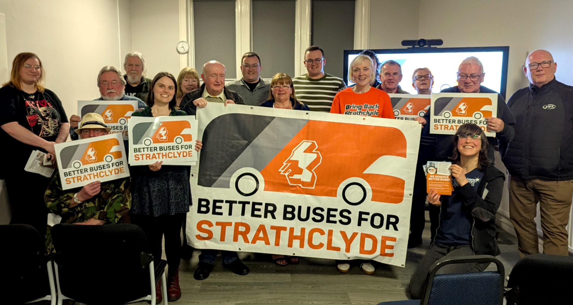 A group of people are holidng 'Better Buses for Strathclyde' campaign signs, in a room with a tv screen behind them.