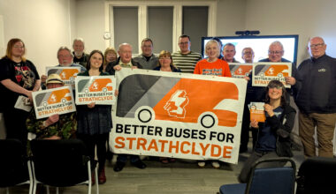 A group of people are holidng 'Better Buses for Strathclyde' campaign signs, in a room with a tv screen behind them.