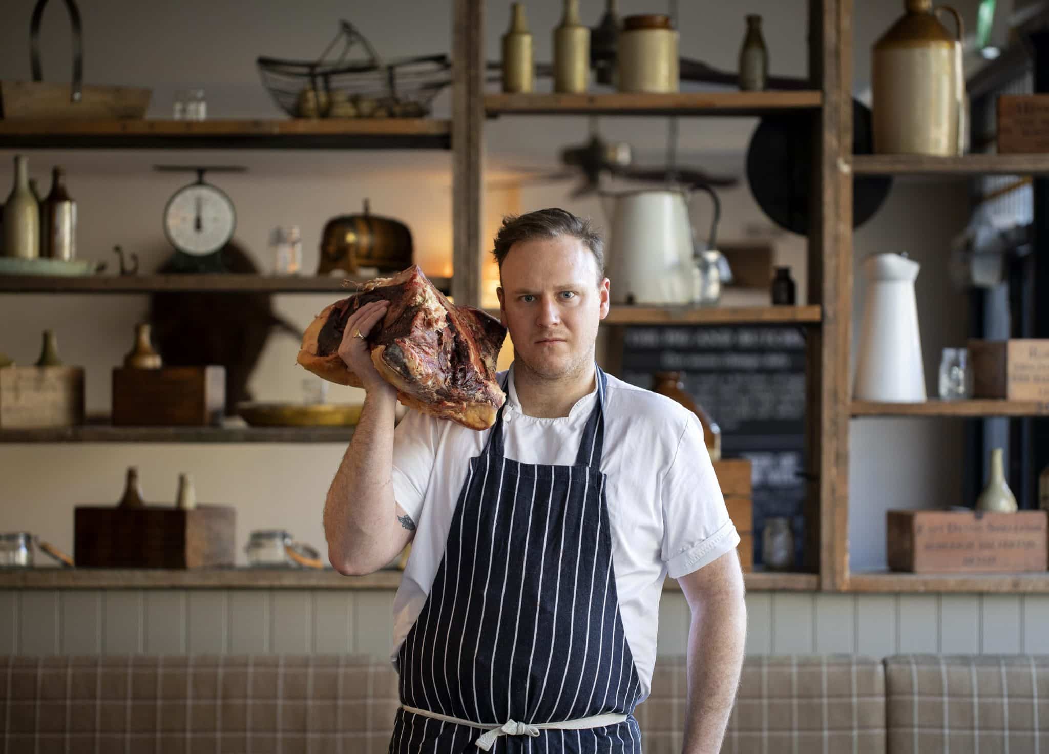 a butcher wearing an apron and holding a big old hunk of beef over his shoulder