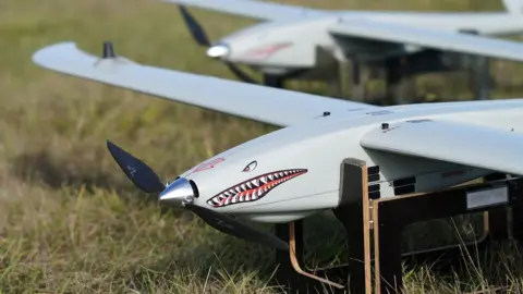 Ukrspecsystems A Ukrspecsystems drone with a shark face and design on its note propped up on a wooden stand in a field.