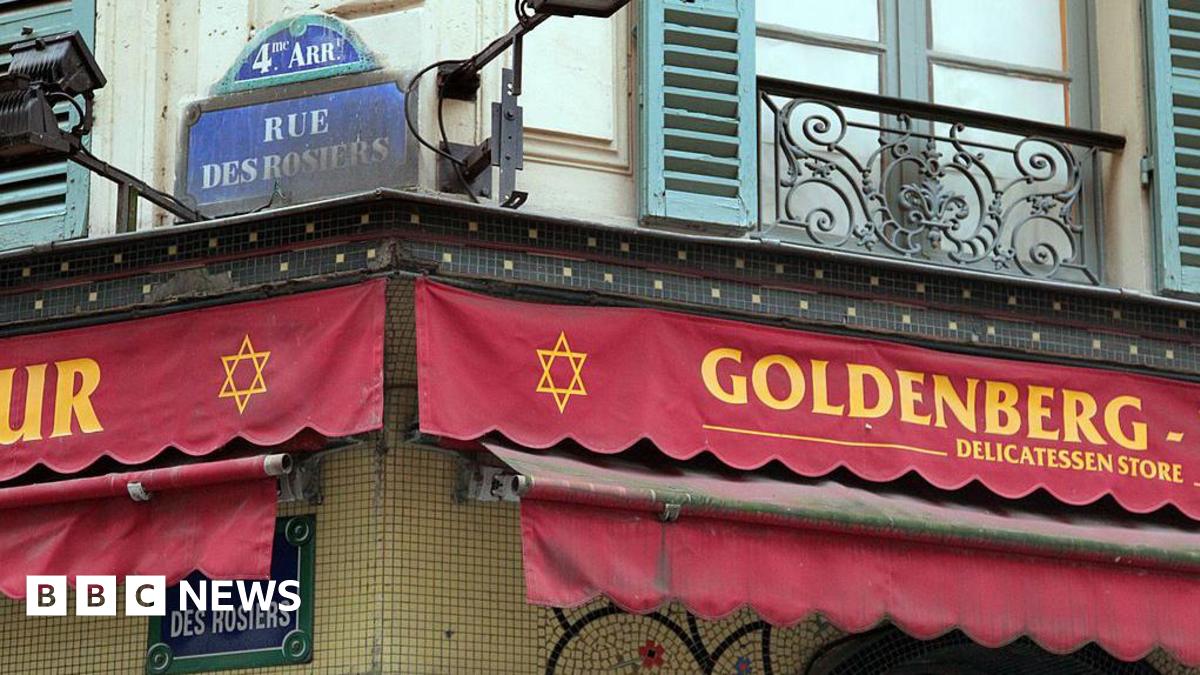 Red scalloped awning advertises Goldenberg's delicatessen stre in yellow font, with the Jewish star of David printed next to the restaurant sign. Above the sign is the street name Rue des Rosiers, 4th arrondisement, in white writing on blue tiling.