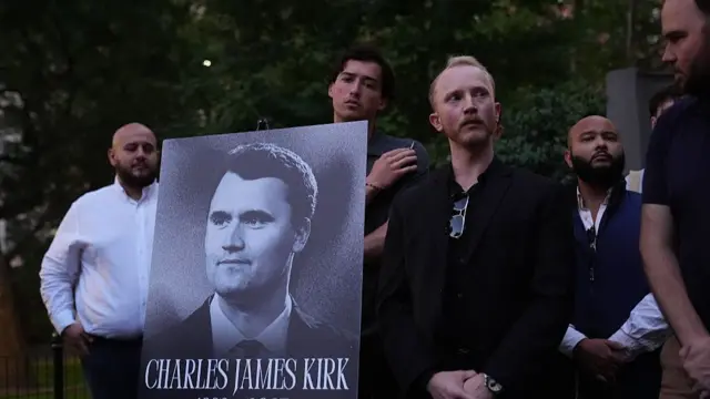 People gathered in mourning carry a large poster with Charlie Kirk's face on it, it says 1993-2025 in black and white.