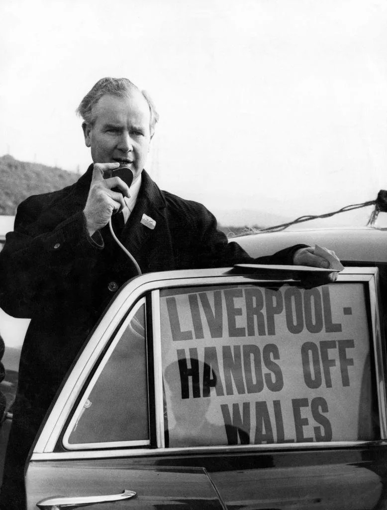 Black and white photograph of Gwynfor Evans. He stands behind an open car door wearing a black long coat, with a welsh flag attached to the lapel collar. He holds a small microphone to his mouth, and a poster can be seen inside the car door which reads: 'Liverpool - hands off Wales'. 