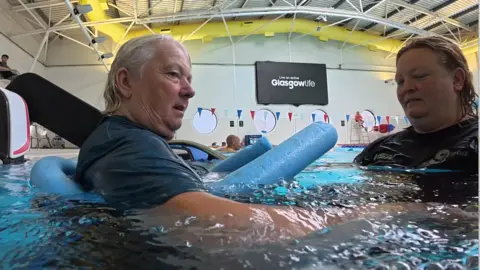 A close up, near the water, of Judith in the swimming pool, a blue pool noodle around her waist, being helped by her swimming coach. The pool walls are decorated with red, white and blue bunting and there is a large "Glasgow Life" banner on the wall.