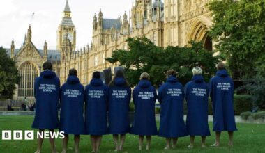 The eight swimmers standing on the green in front of the Houses of Parliament. They are facing away, the slogan 'The Thames Swim Against Water' on the back of their long blue coats.