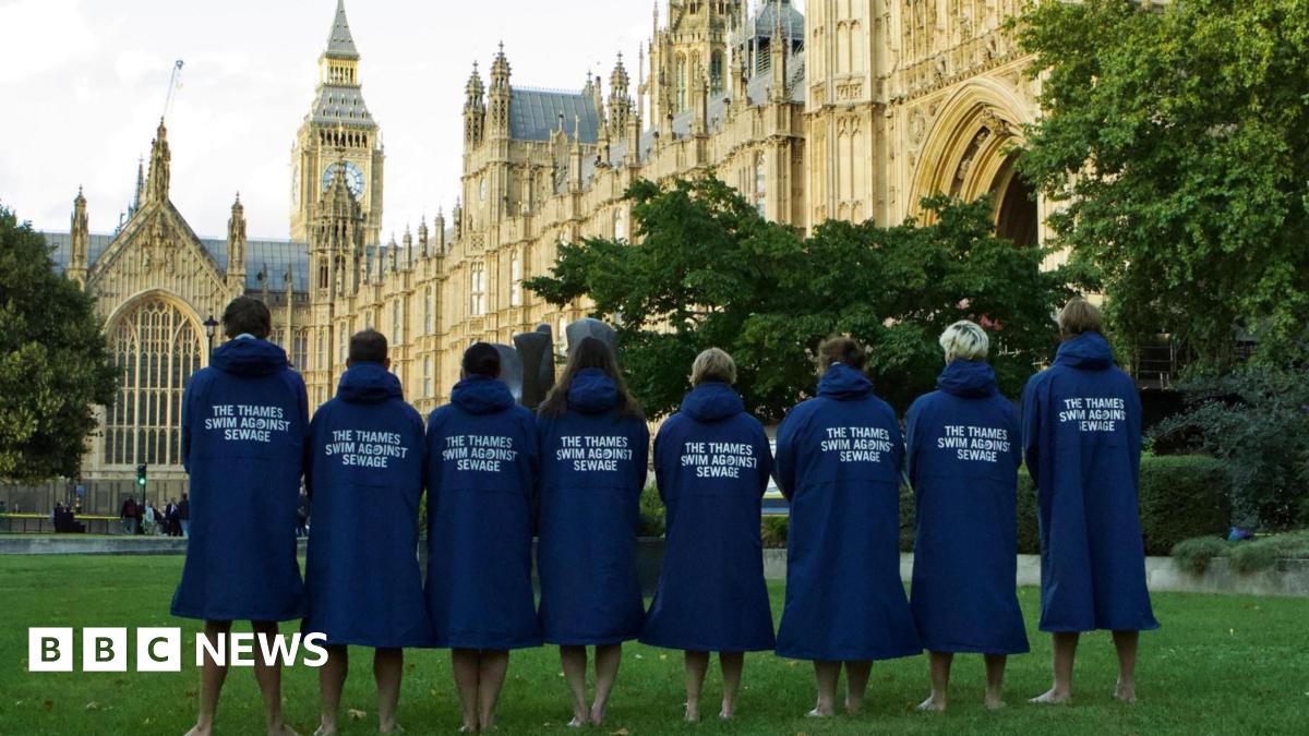 The eight swimmers standing on the green in front of the Houses of Parliament. They are facing away, the slogan 'The Thames Swim Against Water' on the back of their long blue coats.