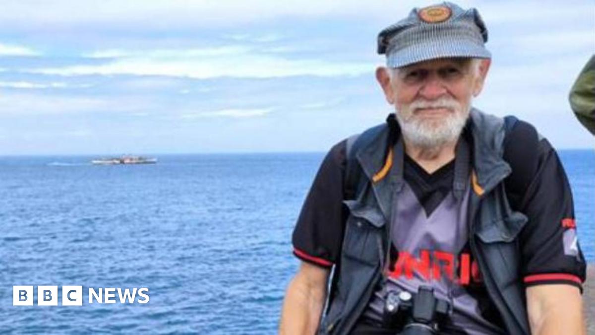 A man with a white beard and a blue hat is seen sitting on a seawall with the ocean behind him. He is wearing a grey jacket a purple shirt and jeans.