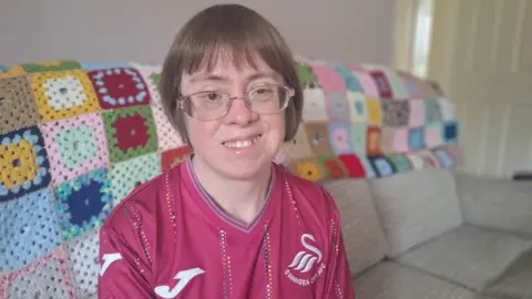 BBC Sara Pickard is sitting on a beige sofa. The sofa has a colourful crocheted blanket thrown over the back of it. Sara is smiling and looking into camera. She has light brown hair in a bob with a fringe and wears glasses. She is wearing a burgundy Swansea City football shirt.