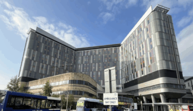 Looking at the many windows of the tall grey hospital building, on a sunny day with blue skies. Several buses and cars are parked in the foreground of the Queen Elizabeth University Hospital in Glasgow