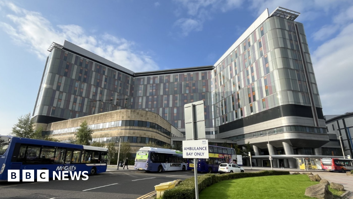Looking at the many windows of the tall grey hospital building, on a sunny day with blue skies. Several buses and cars are parked in the foreground of the Queen Elizabeth University Hospital in Glasgow