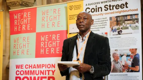 BBC Image shows a man wearing a black suit delivering a speech. 
He is standing in front of two banners about the 'Museum of broken dreams' and the story of the Chapeltown Frontline Self-Build. 