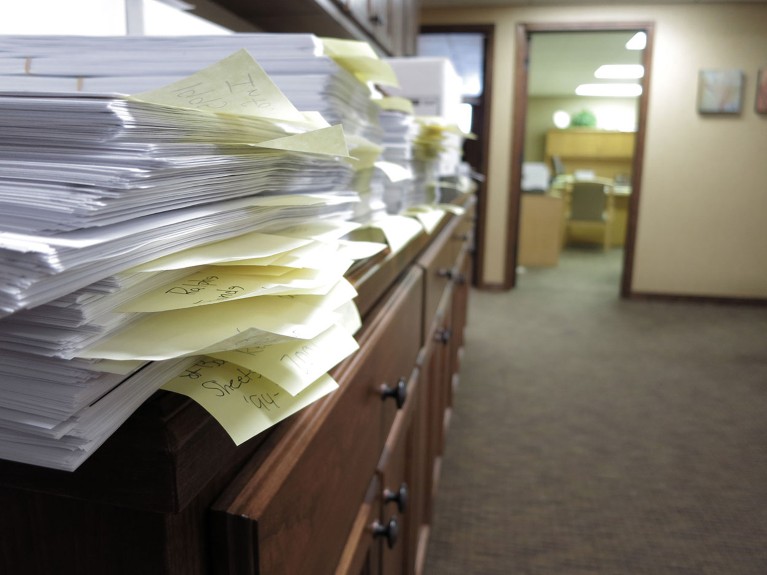 Tall stacks of disorganised paper documents with yellow tabs on a chest of drawers in an empty office.