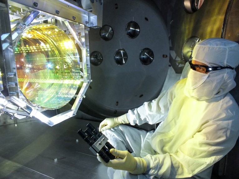A technician wearing an all over white clean room suit looking down a tube with many lights shining of the reflective inner walls