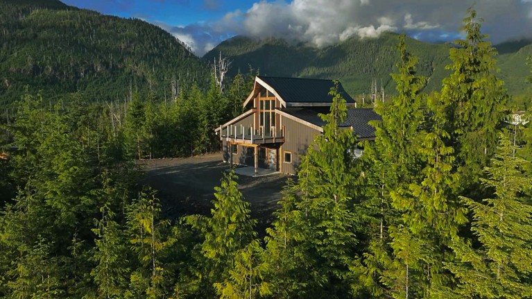 A modern building visible in a clearing of evergreen tress with mountains and clouds in the distance