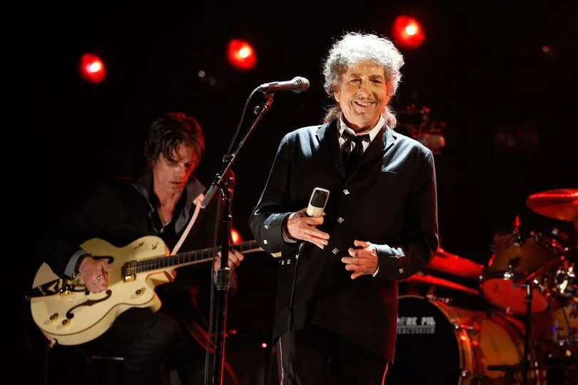 Musician Bob Dylan onstage during the 17th Annual Critics' Choice Movie Awards held at The Hollywood Palladium. He is holding a mic in his hand as a band member plays a cream coloured guitar in the background.