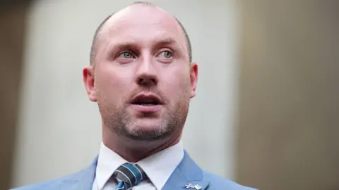 PA Media A man standing, wearing a pale blue suit. He is balding, with stubble. 