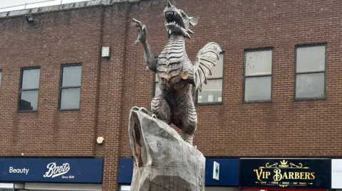 BBC The picture shows the wooden sculpture in Taunton.  The dragon's arm and tongue are missing. There are shops in the background.