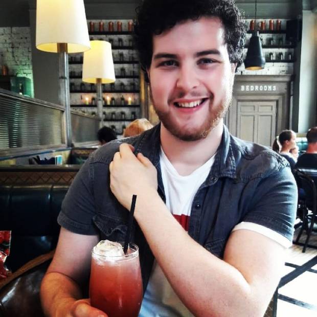 A young man with curly hair and a beard is smiling while holding a drink with ice and whipped cream. He is sitting in a cafe with a lively background.