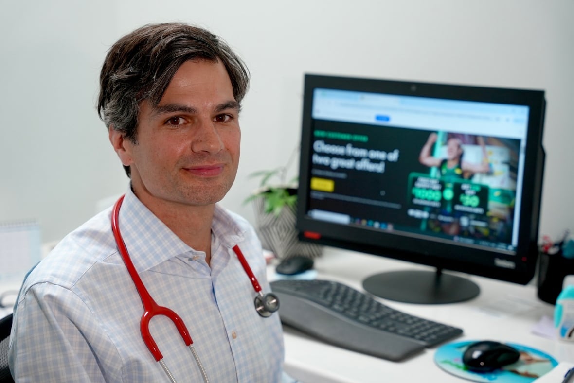 Dr. Kelly sits at a desk. He's wearing a red stethoscope and on his computer in the background, there is a sports betting ad. 