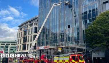 A hydraulic aerial platform is by a tall building. Four fire engines are in front of the building on the image.