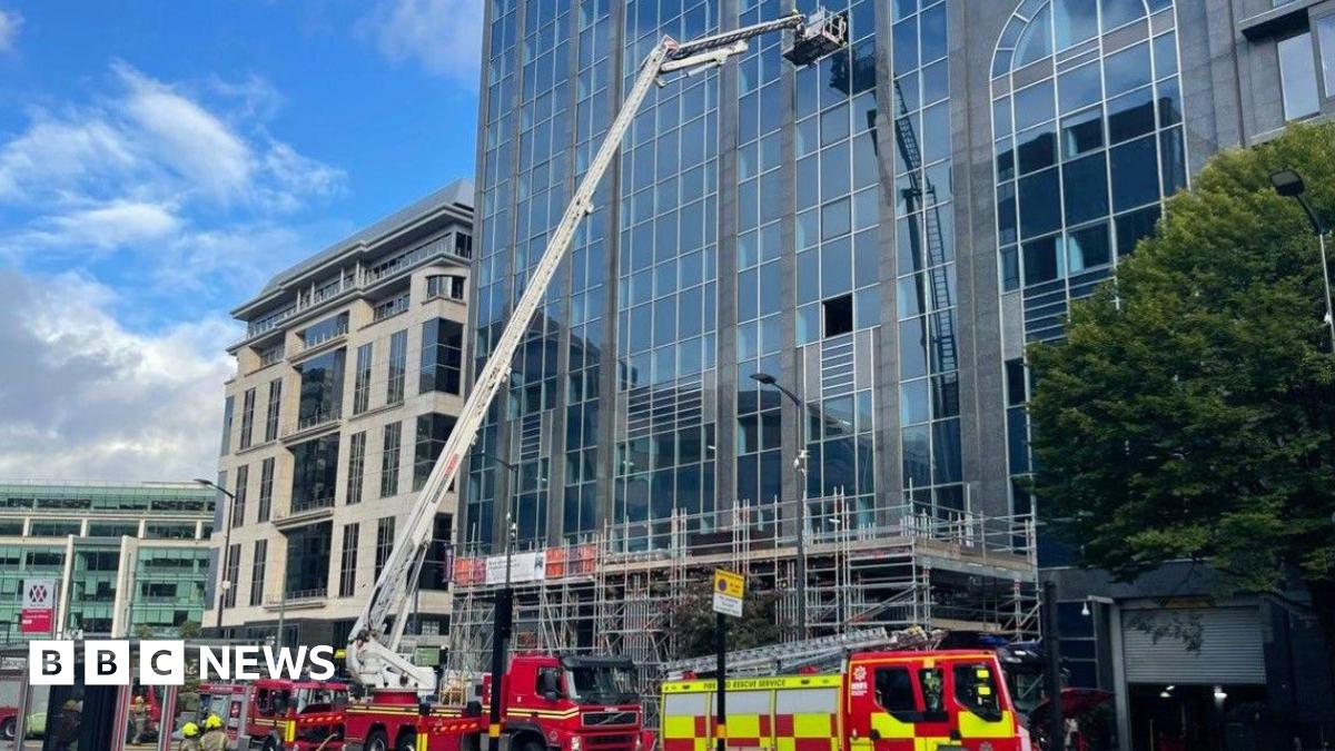 A hydraulic aerial platform is by a tall building. Four fire engines are in front of the building on the image.
