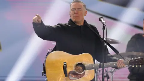 Getty Images Bryan Adams on stage with a drum kit and blue and purple backdrop behind him, and a microphone in front of him. He is pointing towards the audience, has oiled-back hair and is wearing a black shirt.  