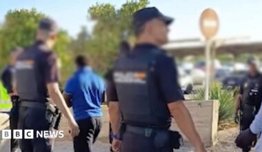 A police handout photo showing the backs of police with their faces blurred, standing next to a coach as people from the migrant boat exit it.
