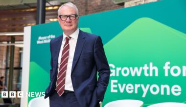 A man with white hair and black glasses. He is wearing a blue suit jacket and trousers with a white shirt and red and white striped tie. He is standing in front off a green board that reads "growth for everyone"