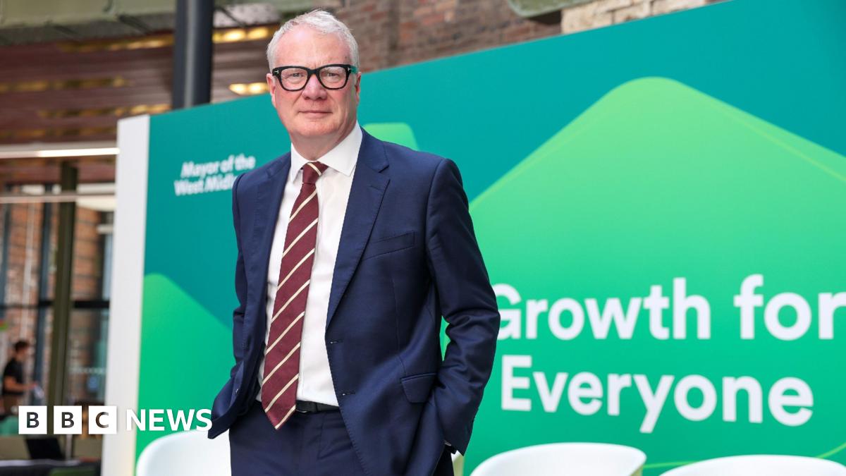 A man with white hair and black glasses. He is wearing a blue suit jacket and trousers with a white shirt and red and white striped tie. He is standing in front off a green board that reads "growth for everyone"