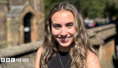 Orla Sonvico smiling at the camera. She has long brown hair and is wearing a black top. There is a wall and an old sandstone building in the distance.