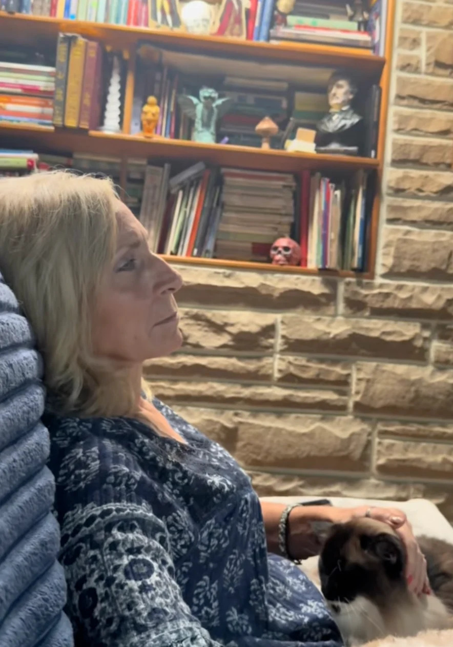 Person sitting on a couch, holding a cat. Books and eclectic decor, including a skull, on shelves behind. Relaxed and thoughtful atmosphere