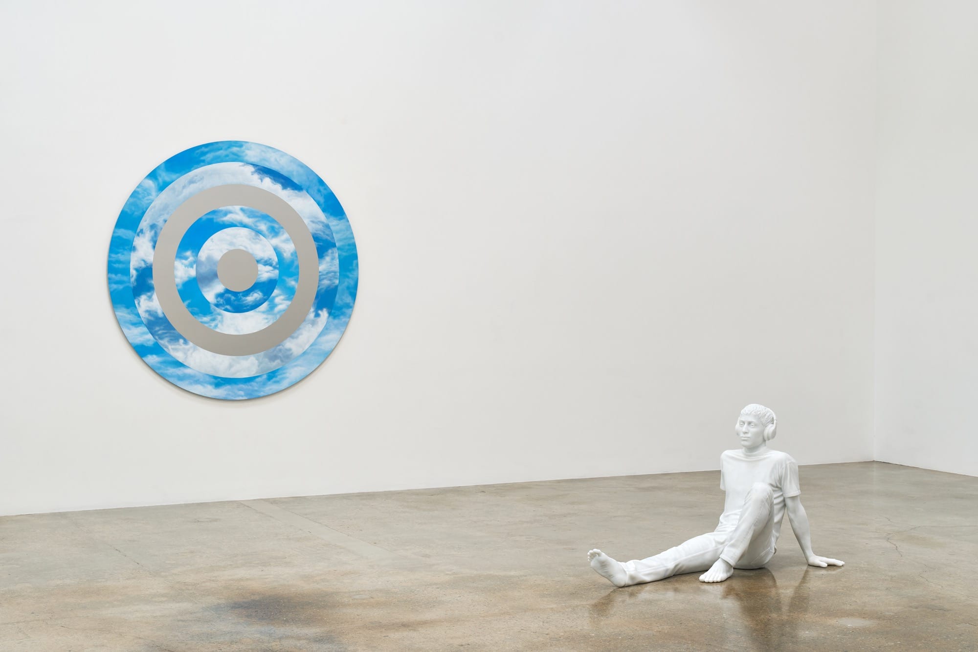 a sculpture by Elmgreen and Dragset of a white figure resting on the floor wearing headphones with a cloudy wall work behind