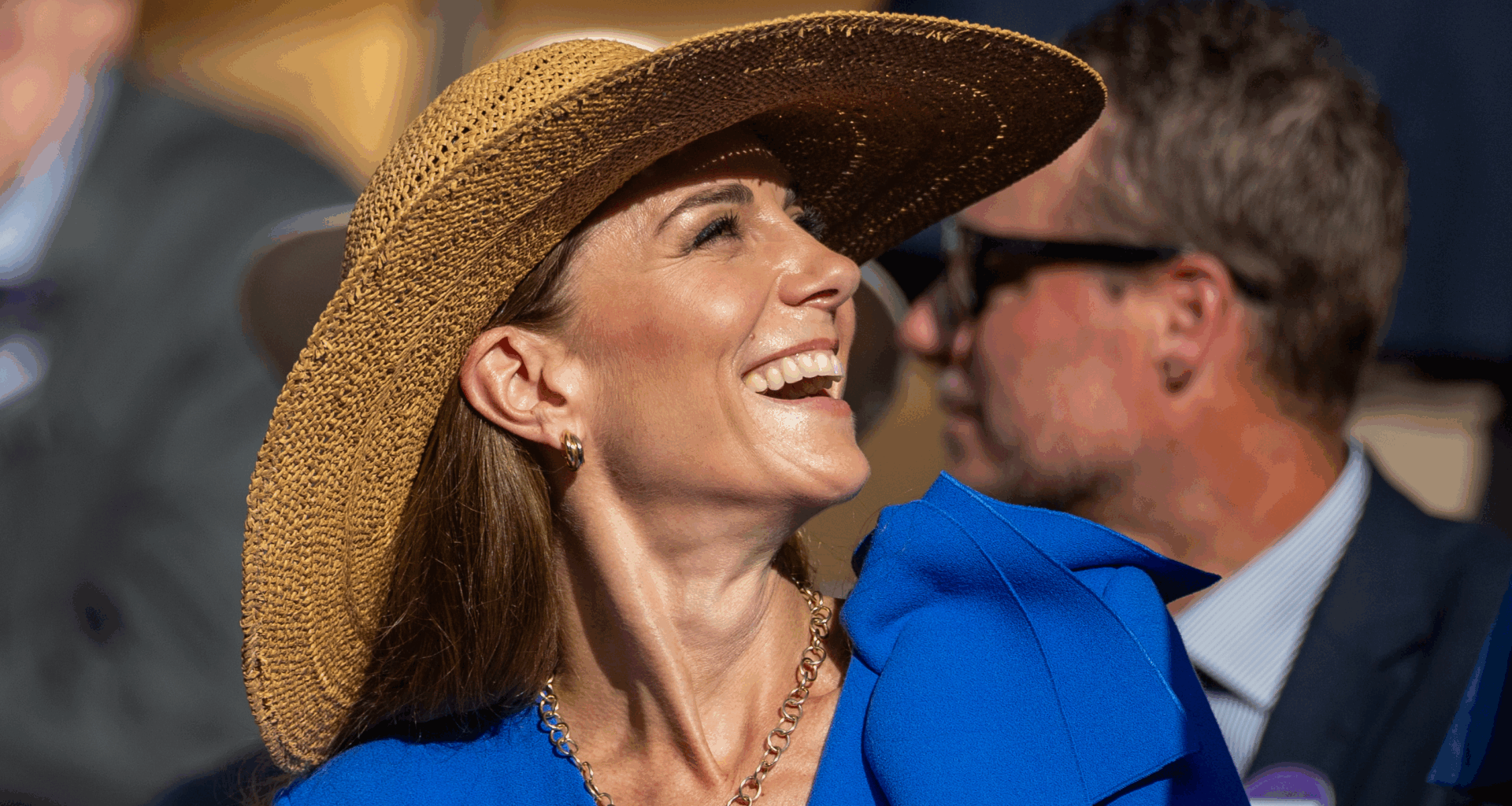 Kate Middleton wearing a blue dress and a straw hat looking up and laughing