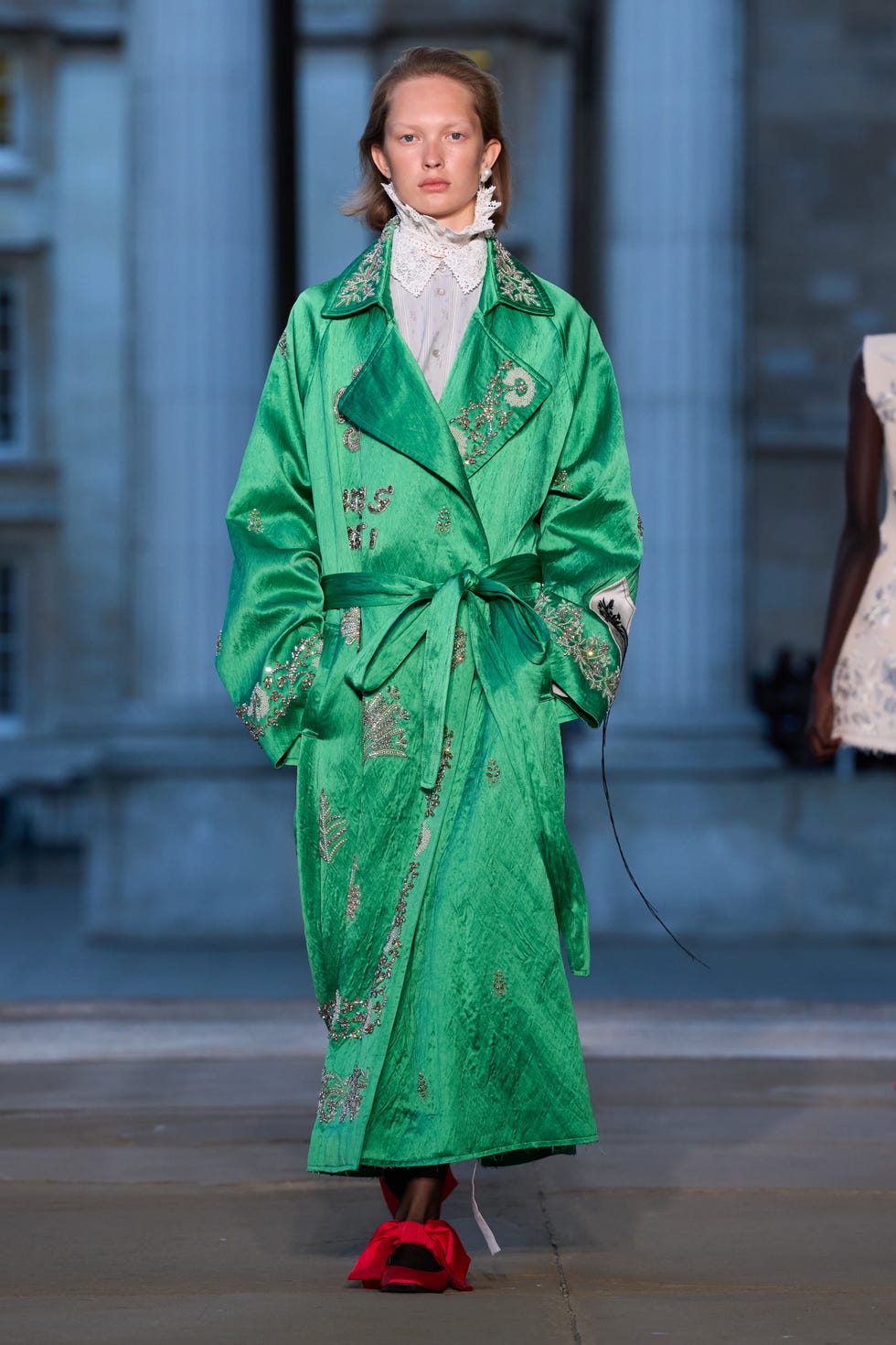 fashion model wearing a vibrant green coat with decorative embroidery and a white highcollared shirt paired with distinct red footwear showcasing a bold runway look