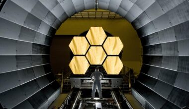 A scientist standing a large metal tunnel with hexagonal gold mirrors at the end of it.
