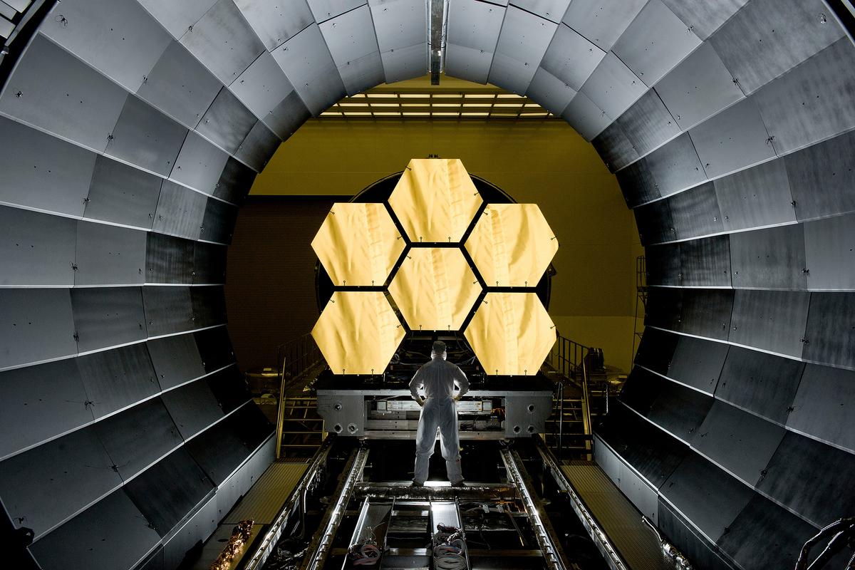 A scientist standing a large metal tunnel with hexagonal gold mirrors at the end of it.