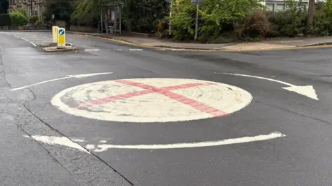 BBC/Louis Reynolds A white mini roundabout with a red cross in the middle. It is painted on a grey tarmac road. There are white arrows around the outside of the circle, and a traffic island can be seen in the background.