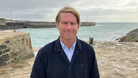 BBC A man with wavy blond hair wearing a blue and white striped shirt and dark blue overshirt standing on a stone harbour with the sea behind him 
