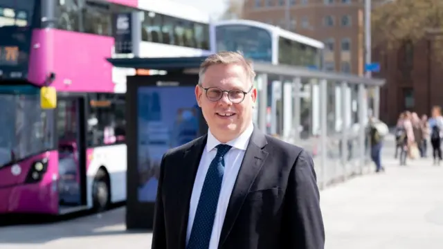 Dough Claringbold wearing a black suit and dark blue tie and round glasses. He is standing in front of a bus stop in Bristol city centre. Behind him there are two buses queuing and several people waiting to board.