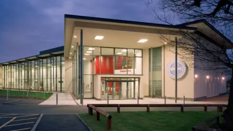 The Royal Orthopaedic Hospital NHS Foundation Trust The entrance to a large white building with lots of black pillars and windows along its side. It is night time and the building is illuminated.
