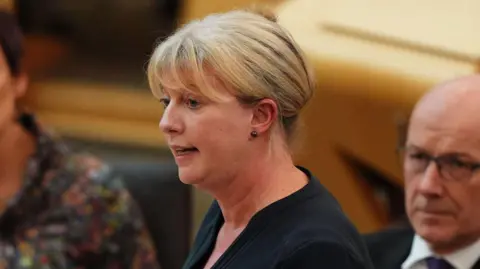 PA Media Shona Robison, a woman with blonde hair tied up, mid speech in the Scottish Parliament chamber. 