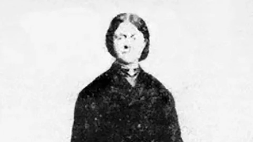 A primitive photograph of Emily outside her Plas Castell home. She is wearing a black coat and holding a hat. Her dark hair is swept back in a bun