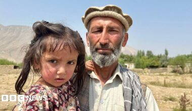 Abdul Wakeel stands looking at the camera while holding his daughter and with his young son standing next to them, against a rural backdrop and blue sky in Shesh Pol in the north-eastern Badakhshan province of Afghanistan.