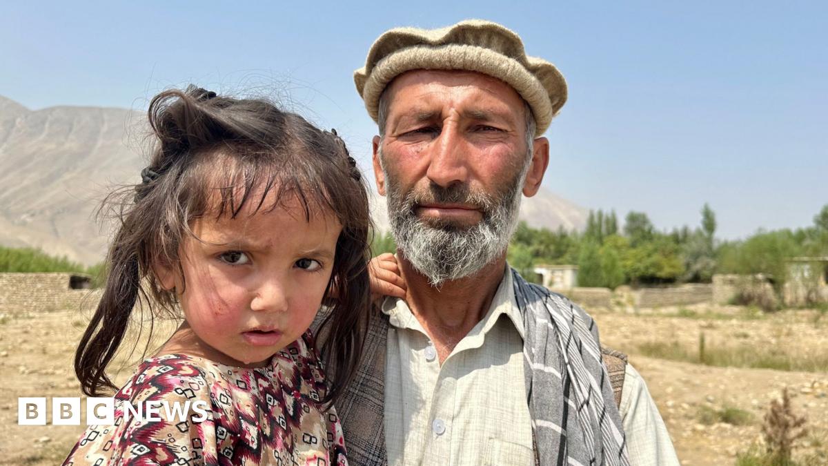 Abdul Wakeel stands looking at the camera while holding his daughter and with his young son standing next to them, against a rural backdrop and blue sky in Shesh Pol in the north-eastern Badakhshan province of Afghanistan.