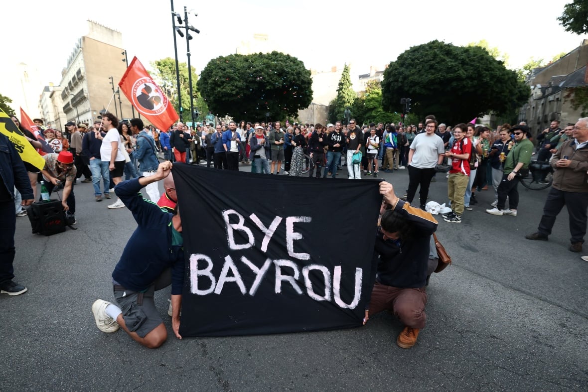 People hold up a black sign with white lettering that reads: Bye Bayrou, as people gather in the street behind them