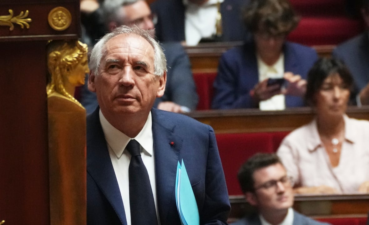 A grey haired man in a blue suit stands looking up to his right in a legislative chamber