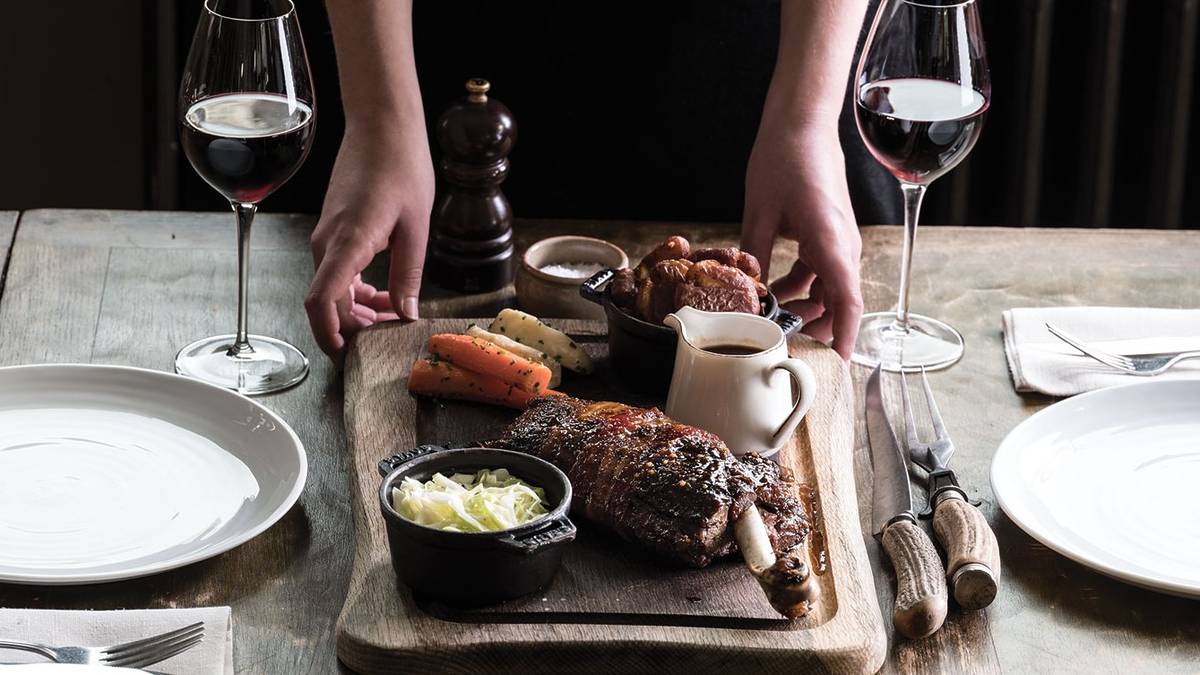 someone setting down a heaving platter containing a beef roast and veg sides, between glasses of wine and plates