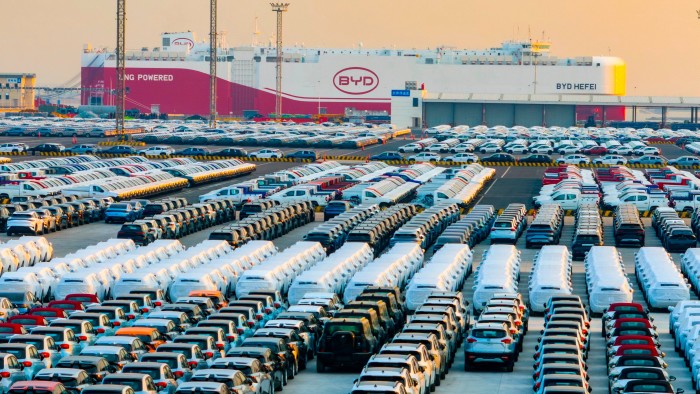 Rows of new energy vehicles are parked at a port terminal, with the BYD Hefei ro-ro ship in the background preparing for export.