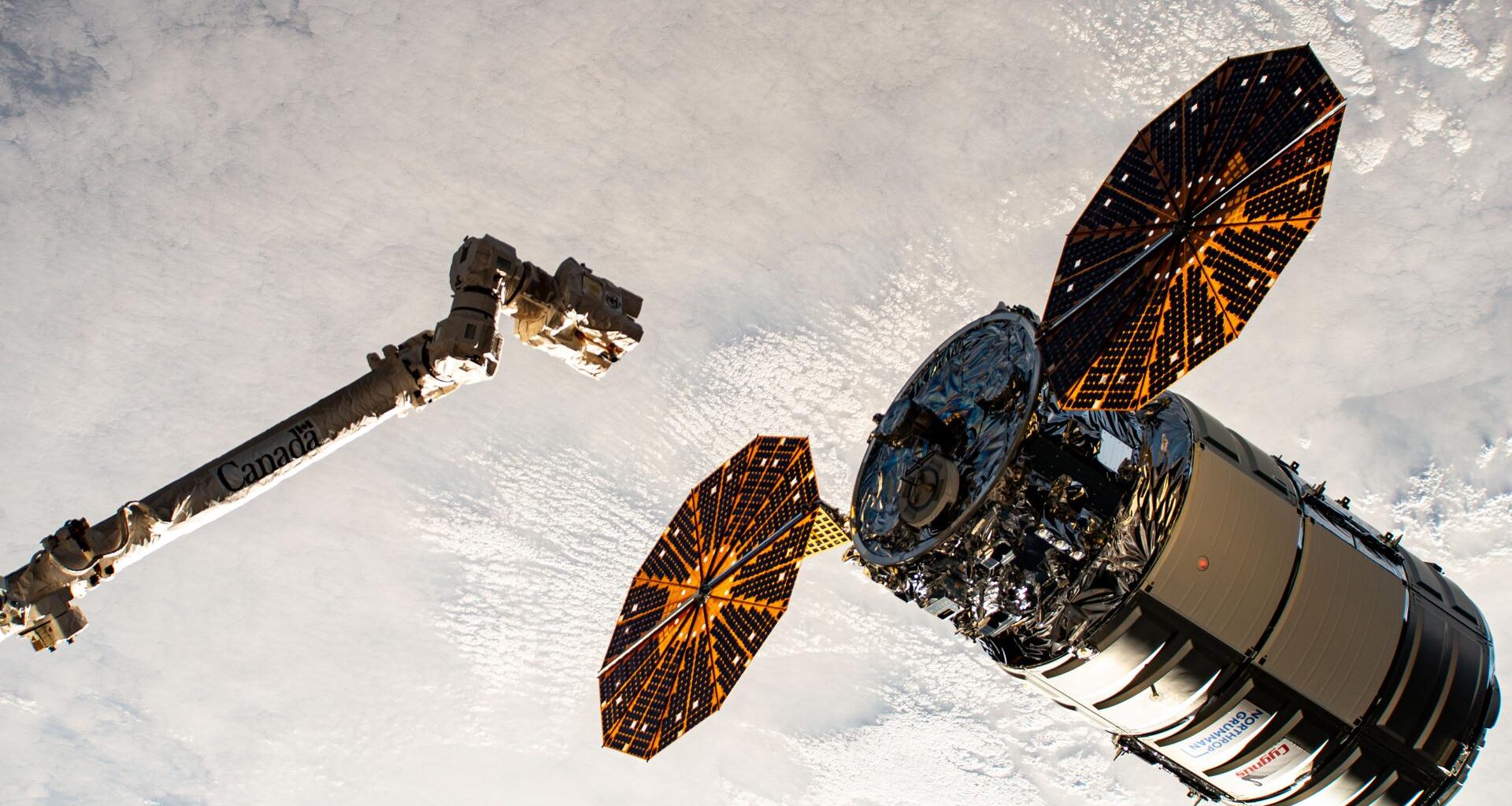 A cylinder-shaped spacecraft with prominent cymbal-shaped solar arrays on either side of it nears a long robotic arm, with a cloudy Earth below.
