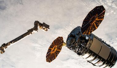 A cylinder-shaped spacecraft with prominent cymbal-shaped solar arrays on either side of it nears a long robotic arm, with a cloudy Earth below.